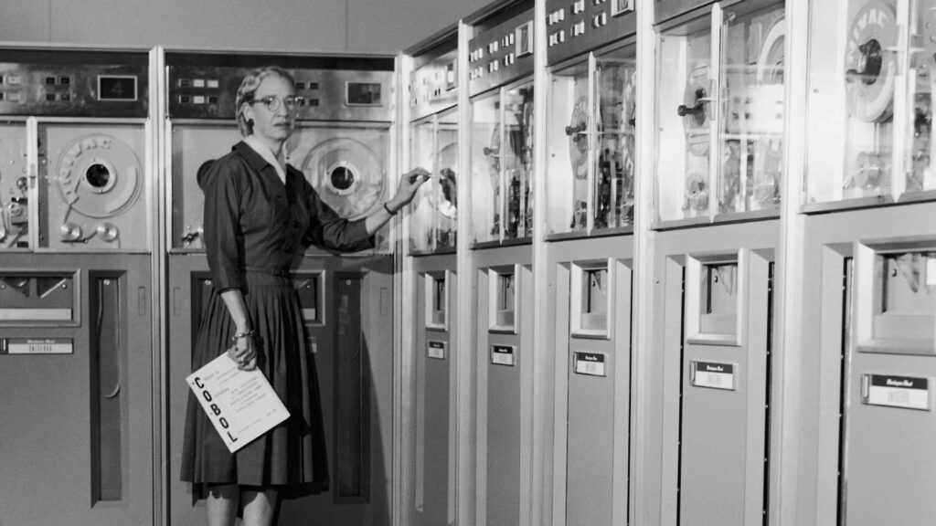 Photo of Grace Hopper standing in front of early computers.